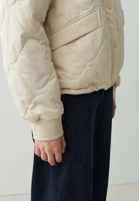 Puff-Jacke in hellbeige mit Steppmuster, gerippten Bündchen, frontaler Tasche und taillierter Passform. Kombiniert mit dunklen Jeanshosen.