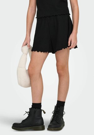 Persona che indossa pantaloncini neri con bordo smerlato e stivali neri stringati, tenendo una borsa bianca testurizzata al proprio fianco.