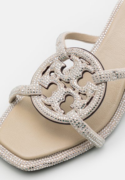 Sandale beige avec une semelle plate, ornée d'accents en strass étincelants et d'un design en découpe circulaire sur la sangle. Semelle intérieure lisse.
