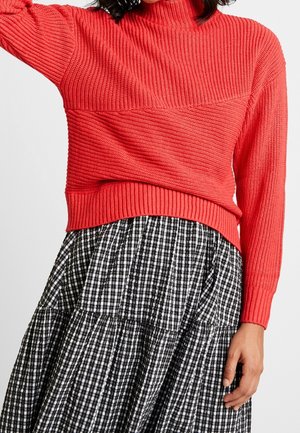 Mujer con un suéter tejido rojo brillante de textura acanalada y una falda a cuadros en blanco y negro.