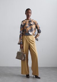 Blouse brune transparente avec motif floral, pantalon large jaune et sac à main gris. La tenue présente des manches évasées et des talons pointus.