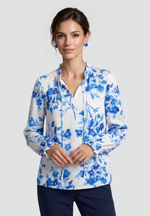 Kvinde iført en hvid bluse med blå blomsterprint, flæsekant og lange ærmer, kombineret med mørkeblå bukser og blå øreringe.