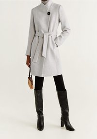 Manteau gris clair en mélange de laine avec un col montant, un bouton noir, une ceinture nouée et des poches latérales, porté avec des leggings noirs et des bottes hautes jusqu'aux genoux.