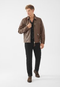 Bruine leren jas met ritsen, zijzakken en een kraag, gedragen over een zwart shirt en gecombineerd met zwarte broek en bruine laarzen.