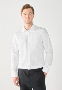 Camicia bianca a maniche lunghe con colletto classico e maniche con polsino. Tessuto liscio con una vestibilità su misura, abbinata a pantaloni scuri.