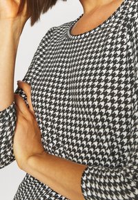 Oděv s černobílým houndstooth vzorem, zaobleným výstřihem a tříčtvrtečními rukávy, vyrobený z texturované látky.