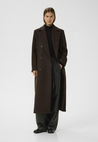 Manteau en laine marron avec un design à double boutonnage, dotée de quatre grands boutons noirs et d'une coupe longue et droite. Associé à un pantalon noir.