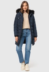 Marineblauwe pufferjas met een met bont gevoerde capuchon, rits aan de voorkant en ribgebreide manchetten, gedragen over een beige gebreide trui en blauwe jeans.