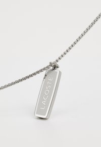 Collier en argent avec un pendentif rectangulaire poli portant le texte gravé "LACOSTE" et une texture lisse.
