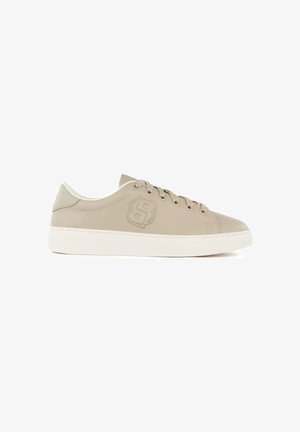 Zapatilla beige de perfil bajo con cordones, suela blanca y un logo en relieve en el lateral.