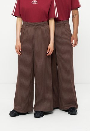 Deux personnes portant un pantalon large marron à fines rayures, des chemises rouges à manches courtes avec des rayures blanches sur les manches, et des chaussures noires, debout côte à côte.