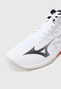 Λευκό αθλητικ�ό παπούτσι με μαύρες πινελιές, λεπτή υφή και λεία επιφάνεια· διαθέτει διακριτικό λογότυπο και κόκκινη λεπτομέρεια στη σόλα.