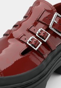 Scarpe in vernice rossa con tre fibbie argentate, ritagli aperti, suola spessa nera e finitura lucida.