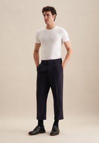 T-shirt blanc ajusté associé à un pantalon plissé bleu marine foncé, avec une coupe droite et des ourlets au-dessus de la cheville. Des chaussures en cuir noires complètent le look.