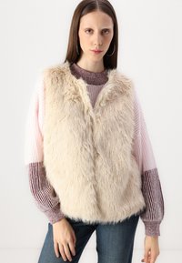 Gilet en fausse fourrure beige avec une texture douce. Porté par-dessus un pull rayé rose et bordeaux, avec des poignets côtelés et une coupe décontractée.