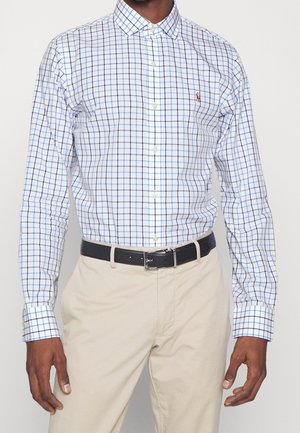 Mężczyzna ubrany w białą koszulę na guziki z długim rękawem w niebieską kratę, beżowe spodnie i czarny pasek, stojący z ramionami swobodnie opuszczonymi wzdłuż ciała.