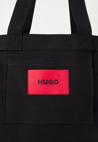 Crna torba od platna s pravokutnom crvenom kožnom oznakom na kojoj piše "HUGO" u crnoj boji. Čvrsta tkanina i minimalistički dizajn.