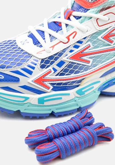 Κοντινό πλάνο ενός μπλε και λευκού αθλητικού παπουτσιού με κόκκινες βέλη, διακοσμημένο δίχτυ και τυρκουάζ σόλα, μαζί με δύο τακτοποιημένα τυλιγμένα κορδόνια σε μπλε και κόκκινο χρώμα.