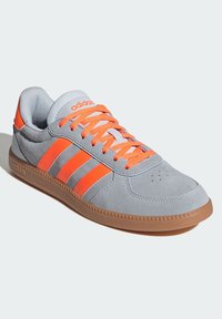 Šedá semišová teniska s oranžovými prvkami, s tromi pruhmi, oranžovými šnúrkami a svetlohnedou gumovou podrážkou. Zaoblený tvar špičky a polstrovaný golier.