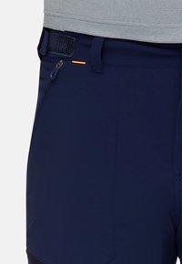 Närbild av marinblåbyxor med zippered ficka och justerbart kardborreband i midjan, bärs med en ljusgrå skjorta.