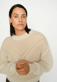 Sweter dzianinowy w jasnobeżowym kolorze, o luźnym kroju, z otwartym wzorem w kształcie litery V i ściągaczami na mankietach. Miękka tekstura z luźnym dekoltem.