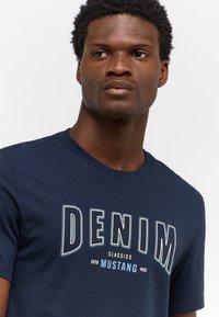 Tamsiai mėlynas medvilninis marškinėliai, papuošti dideliu baltu ir šviesiai mėlynu "DENIM" grafiniu ženklais su tekstu "CLASSICS MUSTANG 1932" po juo.