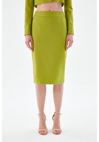 Jupe crayon verte à taille haute, texture lisse, longueur genoux, associée à des sandales à talons aiguilles strappy dans des tons neutres.