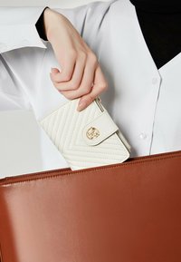Porte-monnaie matelassé blanc avec des accents dorés tenu dans une main, à côté d'un porte-documents en cuir brun, sur un fond clair.