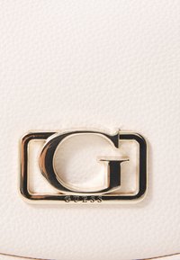 Witte textuurmaterialen bevatten een prominente gouden "G"-logo dat is ingekaderd in een rechthoekig frame, met "GUESS" gegraveerd eronder.