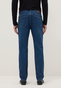 Persona de pie de espaldas, con jeans azules de pierna recta, top negro de manga larga y zapatos negros sobre un fondo liso.