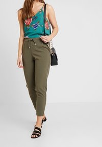 Haut en satin vert floral avec des bretelles fines, associé à un pantalon slim vert olive et des sandales à lanières noires, complété par un petit sac noir.