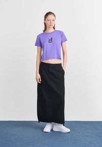 Tricou scurt violet cu un grafic de pisică neagră, asociat cu o fustă lungă neagră și adidași albi, pe o suprafață de covor albastru.