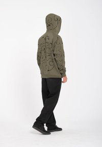 Olijfgroene hoodie met zwart draaiend patroon op de achterkant. Beschikt over geribbelde manchetten en onderkant, gecombineerd met zwarte broeken en schoenen.