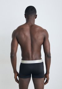 Schwarze Boxershorts mit einem hellgrauen Bund, auf dem "Calvin Klein" in gelber Schrift steht, von hinten auf einem neutralen Hintergrund betrachtet.