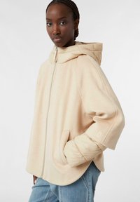 Giacca leggera beige con cappuccio, dotata di maniche trapuntate, cerniera frontale e tasche laterali. Texture morbida e vestibilità comoda.