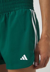 Pantalones cortos deportivos verdes con una cintura elástica, rayas laterales blancas y un logo blanco. Textura suave con elementos de diseño casual.