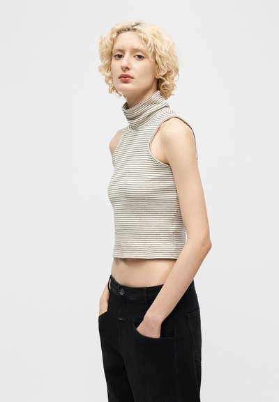 Νεαρό άτομο με κοντά σγουρά ξανθά μαλλιά, φοράει αμάνικη μπεζ και μαύρη ριγέ μπλούζα με ζιβάγκο και μαύρο παντελόνι, με τα χέρια στις τσέπες.