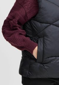 Crni prsluk od puffer materijala s dijamantnim quilt uzorkom, s bočnom džepom i rebrastim rukavima od maroon džempera s teksturiranim pletenjem.