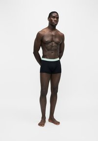 Homme musclé à la peau foncée, debout pieds nus, mains dans le dos, portant un boxer noir avec une ceinture de couleur claire.