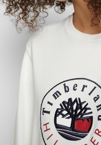 Personne aux cheveux bouclés portant un sweat-shirt blanc avec un grand logo Timberland, représentant un arbre et du texte en noir et rouge.