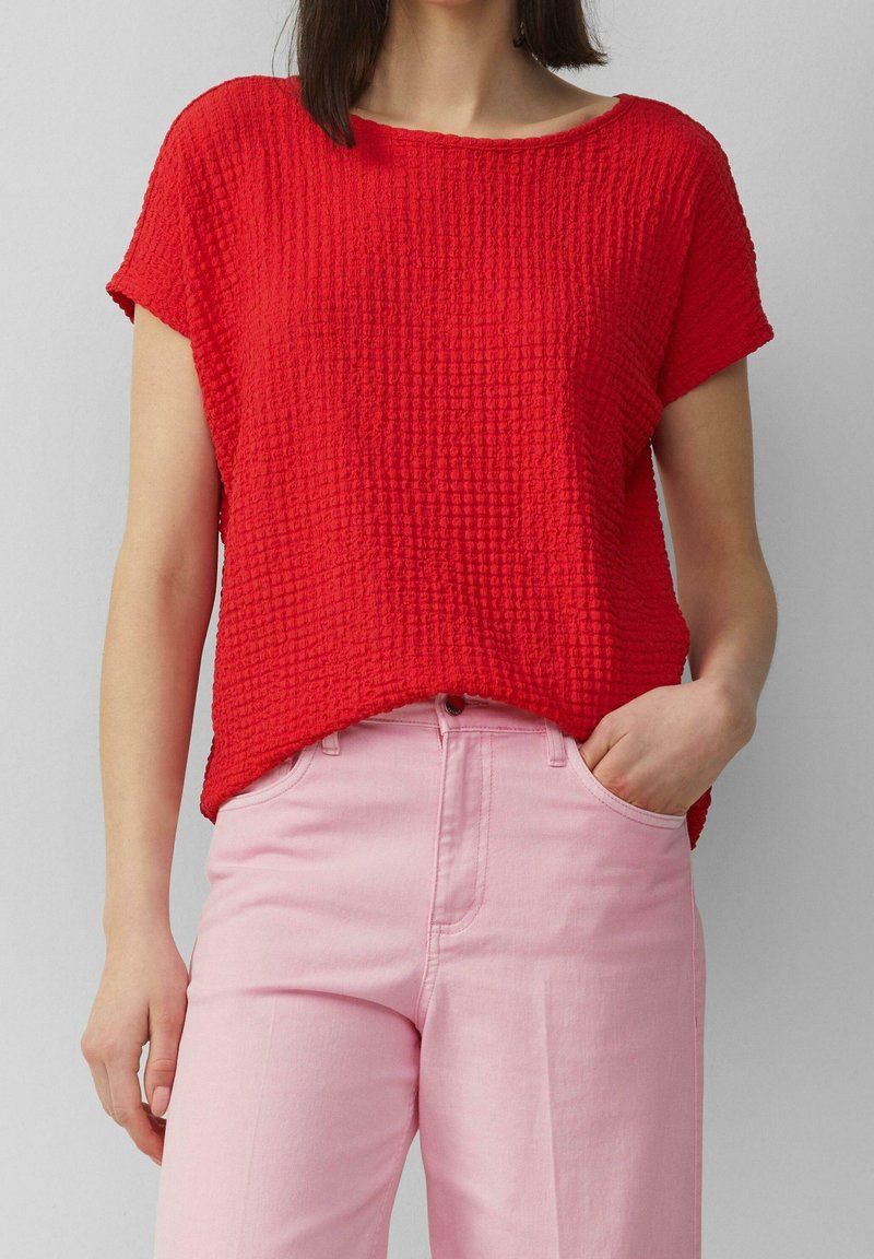 Femme portant un haut rouge texturé à manches courtes et un pantalon taille haute rose clair, une main dans la poche, sur un fond gris uni.