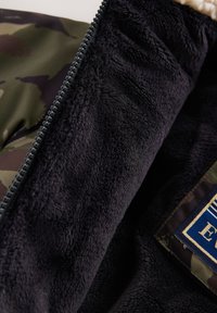 Interior de chaqueta con patrón de camuflaje, forro de felpa suave azul marino oscuro, abertura con cremallera y etiqueta de tela con detalles en azul y dorado.