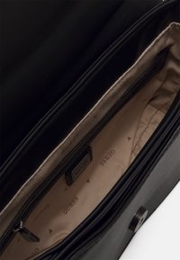 Sort håndtaske med en glat læderoverflade. Indvendigt har den en blød tan-farvet foring med et gentaget "Guess" logo mønster og en lynlåslomme.