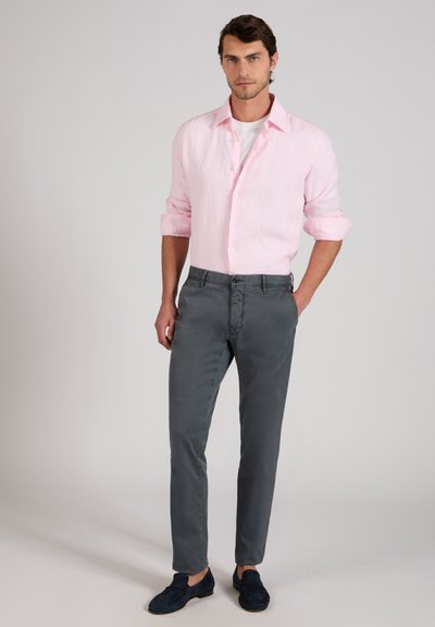 Uomo che indossa una camicia rosa chiaro abbottonata sopra una t-shirt bianca, pantaloni grigi e mocassini scuri, in piedi con una mano in tasca.