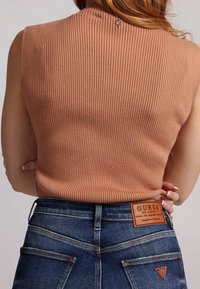 Top a coste senza maniche di colore pesco tenue, abbinato a jeans attillati blu scuro. Notabile applicazione in pelle con logo sulla tasca posteriore dei jeans.