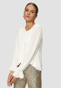 Weißes, gefaltetes Blusenshirt mit langen Ärmeln und Rüschenbündchen, kombiniert mit hoch taillierten, schimmernden goldenen Pailletten-Hosen. Schlichte Dekolleté-Gestaltung.