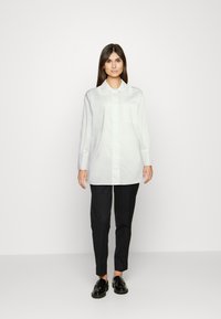 Camisa branca de botões com um grande colarinho, mangas longas e um bolso no peito; combinada com calças pretas e sapatos pretos.