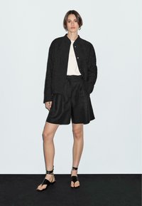 Ung kvinna som står, iklädd en svart texturerad jacka, vit topp, svarta vida shorts och svarta sandaler med ankelsnodd mot en enkel bakgrund.