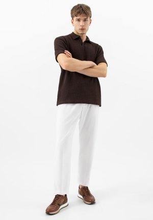 Junger Mann mit verschränkten Armen, der ein schwarzes, kurzärmeliges, strukturiertes Polohemd, weiße Hose und braune Turnschuhe vor einem weißen Hintergrund trägt.