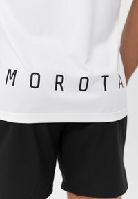 Hvid atletisk skjorte med sort tekst "MOROTA" på ryggen, parret med sorte shorts, der har en glat tekstur og atletisk pasform.